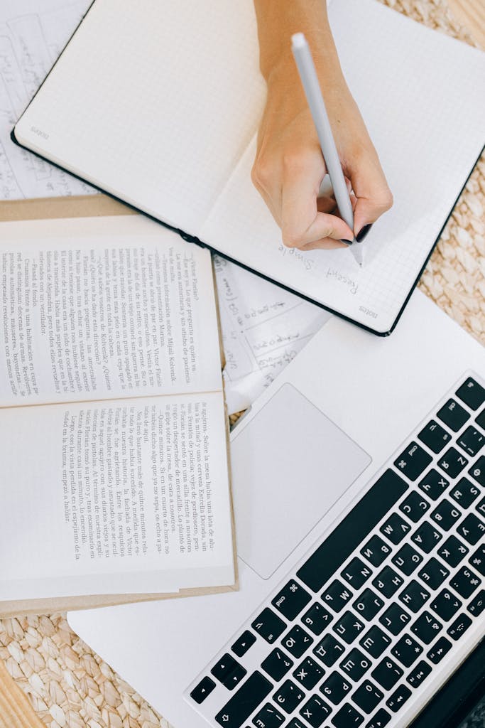 Close-up of a woman's hand writing in an open notebook on a desk with a laptop and books.